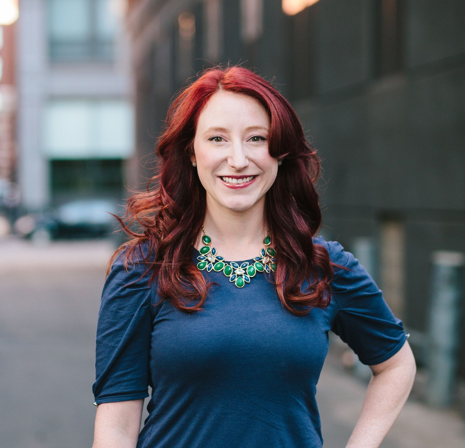 A headshot of Jen smiling outside while wearing a blue shirt and green necklace. 