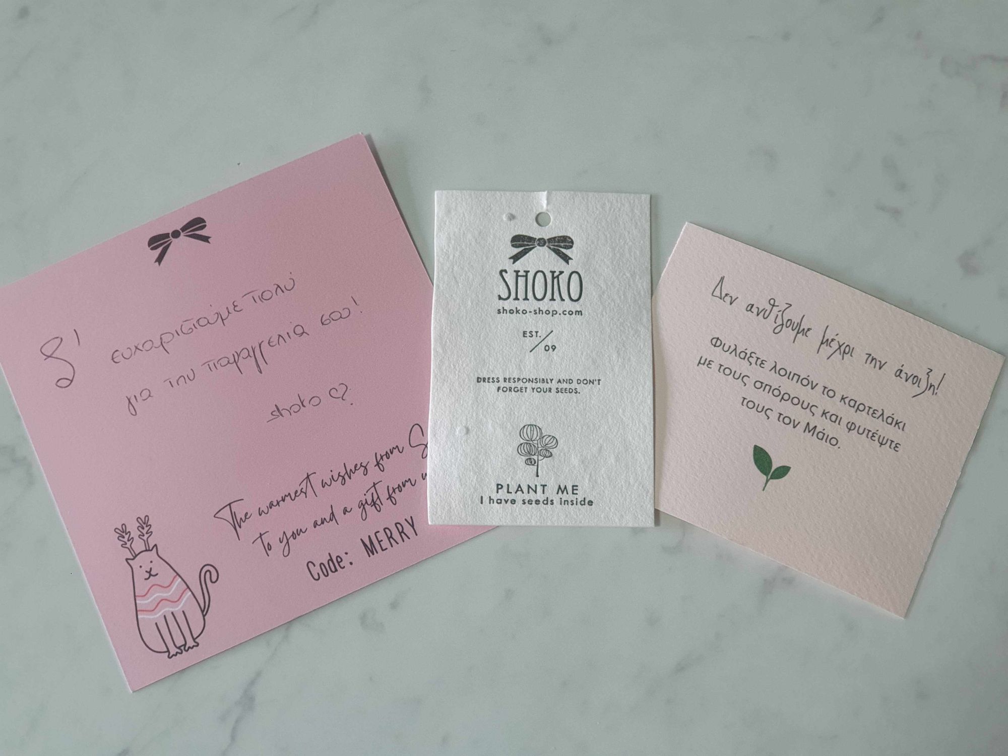 Sustainable brands–An image of three notes from Shoko that were included in an order. They are on pink and white branded pieces of paper with hand-written messages in Greek. The tag in the middle is white and has the Shoko logo couples with an image of a tree and the message: Plant Me. I have seeds inside. 