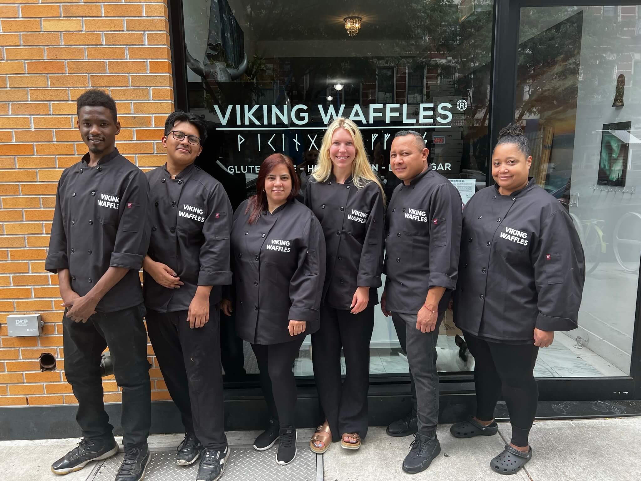 An image of 6 Viking Waffles employees standing in a line, smiling outside their bakery with “Viking Waffles” printed on the window. They are all dressed in black pants and black chef jackets, with the brand logo printed on them. The founder, Benedicte Engen is a tall blonde woman and is standing in the middle. 