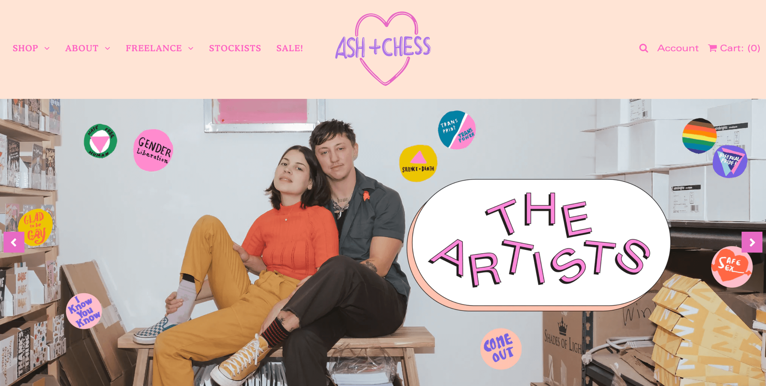 A screenshot from the Ash + Chess homepage. The image shows an image of Ash and Chess entwined on a bench together in their studio. The title is in a retro, bright color font and says The Artists and there are various graphic icons overlaying the image with LGBTQIA+ positive messages such as: come out, I know you know, glad to be gay, gender liberation, and more.