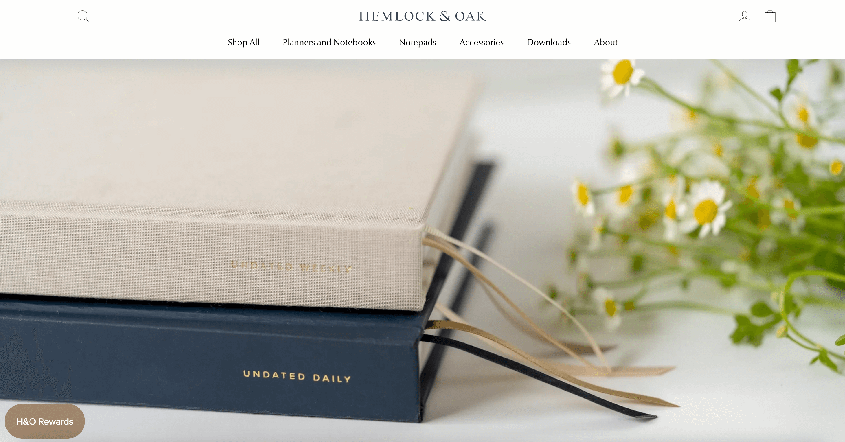 A screenshot from Hemlock & Oak’s homepage showing a photo of 2 of its undated daily journals on a white desk with some meadow flowers surrounding them. 
