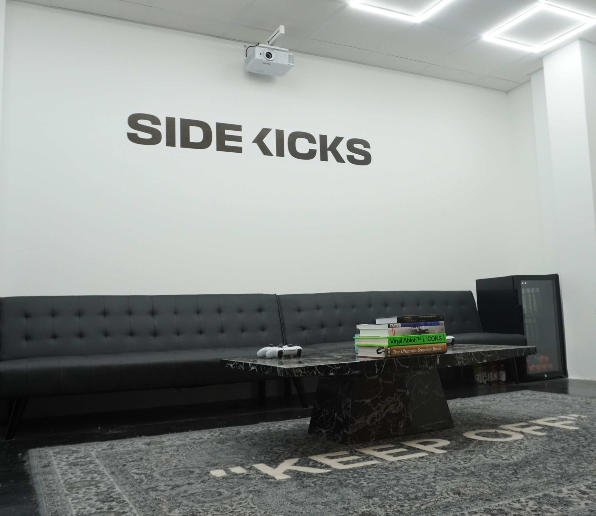 An image of the Side Kicks VIP Room. The logo is printed on the wall behind a chic black couch. There is a coffee table with several books, a video game controller, and a fully stocked mini fridge.